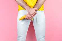 Potency And Men's Health. A Man In White Jeans, Legs Apart, Holds A Banana Near The Genitals. Pink Background. Close Up Of Hands