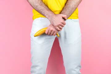Potency and men's health. A man in white jeans, legs apart, holds a banana near the genitals. Pink background. Close up of hands