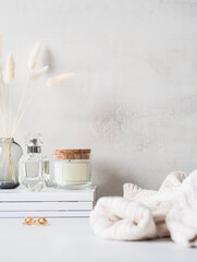 Modern feminine still life of white dry lagurus flower in glass vase, glass scented candle and a bottle of perfume on stack of white magazines on light background. Front view. Copy space