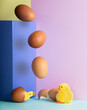 Staged joke setting of eggs falling from a ledge and two artificial chicks emerging from broken shells at the base, with colour theme of pale magenta cyan and yellow