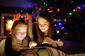 Wall Mural - Two cute young sisters using a tablet pc at home by a fireplace in warm and cozy living room on Christmas eve. Winter evening with family and kids.