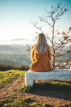 Woman On A Yellow Coat Sitting On A Bench Contemplating The Landscape