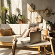 © Floral Deco - Boho style modern home interior design. Laptop, sofa, pillows, home plants, carpet and decorations against concrete wall. Bohemian sitting room with warm sun light shadows on the wall.