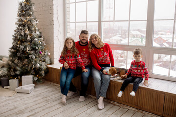 Wall Mural - Young Caucasian beautiful family mom and dad are sitting embracing with their son, daughter and dog on the windowsill of the house among the Christmas decorations. Family portrait at home
