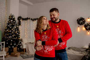 Wall Mural - Young caucasian couple man and woman hugging and laughing while standing at home in living room with christmas tree
