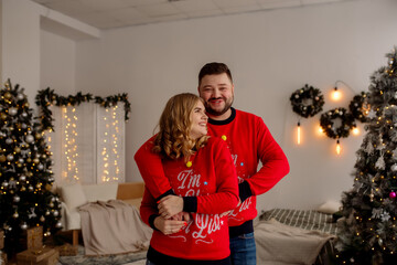 Wall Mural - Young caucasian couple man and woman hugging and laughing while standing at home in living room with christmas tree
