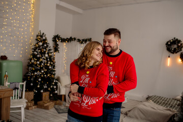 Wall Mural - Young caucasian couple man and woman hugging and laughing while standing at home in living room with christmas tree
