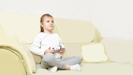 little girl sits in the living room and plays video games on a console or PC with a joystick, looking at a screen or TV in interior room at home. playing online video games having fun. 