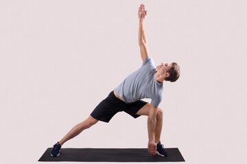 Wall Mural - Strong young man standing in yoga pose on sports mat over light studio background, full length portrait