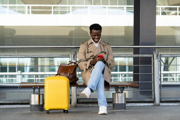 Smiling Afro-American traveler man with yellow suitcase sitting on bench in airport terminal or railway station, using mobile phone, calling taxi, waiting public transport.  