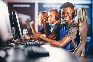 Side view of a cheerful african guy, professional cybersport gamer wearing headphones looking at camera and smiling while participating with team in eSport tournament