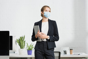  executive in medical mask with laptop looking away, while standing near workplace in office