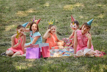 Little Girls On A Picnic Celebrate Their Birthday In Carnival Masks. Girls In Bright Dresses. The Bright Green Grass.