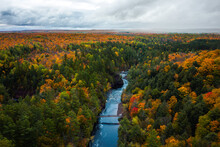 Beautiful Aerial Above The Bad River And A Pedestrian Foot Bridge At Copper Falls With Colorful Fall Foliage Lining The River Banks And Cloudy Sky Above The Horizon In Autumn In Mellen, Wisconsin.