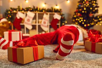 Gift box tied with red ribbon beside tired drunk Santa lying asleep on floor in living room