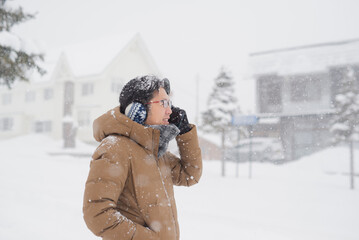 Asian man in glasses using  smartphone on city street during snowfall in winter.