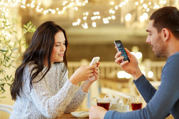 Wall Mural - people, communication and dating concept - happy couple with smartphones drinking tea at cafe or restaurant over festive lights background