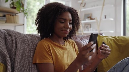 Poster - Smiling young african woman holding credit card and smartphone sitting on couch at home