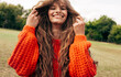 © iuricazac - Positive young woman playing with her long red hair wearing an orange knitted sweater posing on the nature background. Pretty female playing with her hair outdoors in the park.