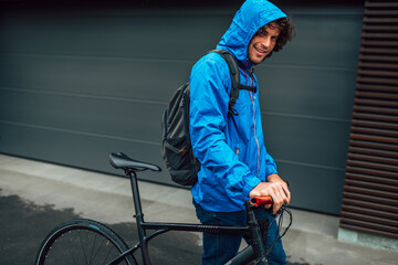 Handsome man in a blue raincoat posing with his bike before bicycling on a rainy day next to the house. Male courier with curly hair delivers parcel cycling with a bicycle.