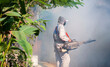 Outdoor healthcare worker using fogging machine spraying chemical to eliminate mosquitoes and prevent dengue fever in the midst of many chemical fumes on overgrown at slum area 