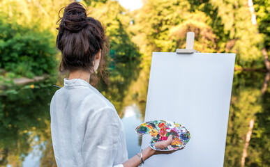 girl artist summer park pond, background forest water shore trees, draws picture, view front, hand palette with paints easel. Clothes white shirt. White blank canvas looking at landscape.