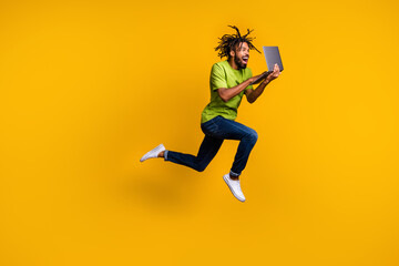 Full length photo portrait of excited programmer with dreadlocks jumping up holding laptop in hands isolated on vivid yellow colored background