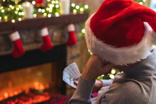 Sad Man In A Santa Claus Hat Thinking Counting American Dollars Sitting Near Christmas Tree And Fireplace. Spending Money On Gift At Christmas Time. Concept.