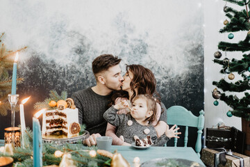 Wall Mural - Young family with a daughter in festive outfits at a served table with candles, garlands, sparklers and a cake near the Christmas tree on New Years Eve