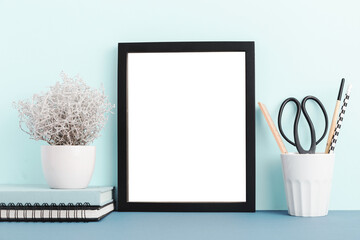 Wall Mural - Table with blank picture frame or poster, desk objects, office supplies, books and plant on a blue background. Mock up.	