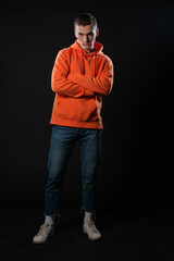 Wall Mural - Young handsome man in jeans and orange hoodie posing while standing on a black background.