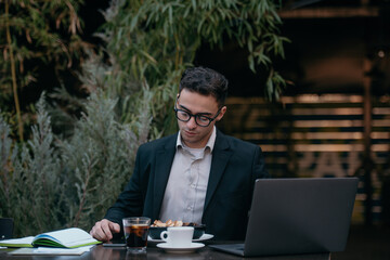 Young successful businessman working on a laptop while sitting in coffee bar during work break lunch. Speaking on his phone while working and drinking coffee.	