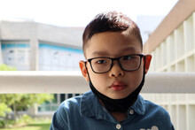 A Portrait Photo Of Asian Young Short Hair Boy Kid Wearing Glasses , Black Cloth Mask And Blue Shirt With Background Of School Building During Waiting For The Parents To Go Back Home