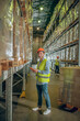 © zinkevych - Young warehouse worker in helmet standing near containers and holding a tablet