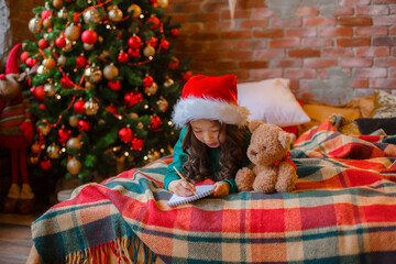 Wall Mural - little Asian girl in pajamas near the Christmas tree writes a letter to Santa Claus Christmas, new year