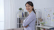 © PRPicturesProduction - confident asian female manager in professional blazer smiling facing camera is standing with arms crossed in modern office.