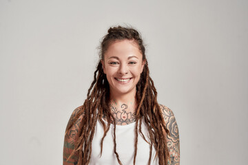 Wall Mural - Portrait of young tattooed caucasian woman with dreadlocks wearing white shirt, smiling at camera while posing isolated over grey background