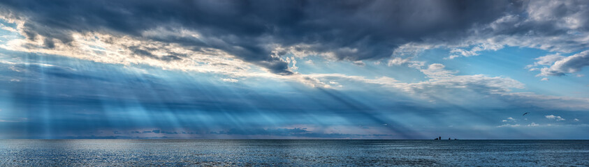 Sun rays through dark clouds over the Black sea with small rocks on the horizon, horizontal panorama, high resolution