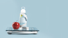 Red apple, white tape measure around a lemon water glass bottle on top of glass bathroom scale on blue. Front view copy space. Healthy lifestyle, weight losing management and hydration concept