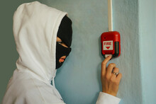 School Girl Pulling Fire Alarm False Signal On The Wall Next To The Door. Girl Wearing Black Mask And Hoody.