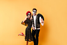Funny Shot Of Full-length Couple Of Lovers In Halloween Costumes Having Fun Against Backdrop Of Orange Wall.