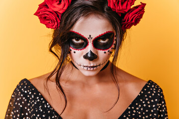 Wall Mural - Close-up photo of brown-eyed woman with carnival face art. Mexican model is angry looking at camera on orange background