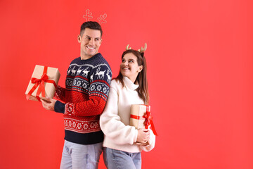 Wall Mural - Beautiful happy couple in Christmas headbands and sweaters holding gifts on red background. Space for text