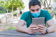 © Marcela Ruty Romero - Young man with a protective mask watching multimedia content on a tablet, sitting in a park.
