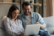 © fizkes - Happy millennial man and woman sit on sofa relaxing watch funny video on laptop laughing together. Smiling young Caucasian couple have fun rest look at computer screen enjoy online shopping from home.