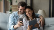 © fizkes - Wide banner panoramic view of happy millennial spouses man and woman relax on sofa using modern gadgets together. Smiling Caucasian couple rest at home have fun browsing electronic devices.