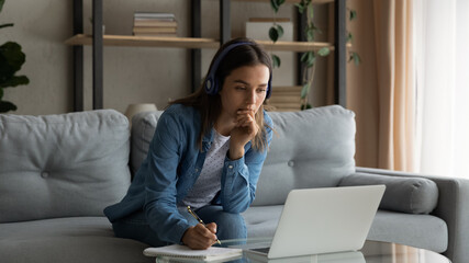 Wide banner panorama view of serious millennial female student on headphones study distant on laptop from home. Focused young woman in earphones look at computer screen working online on device.