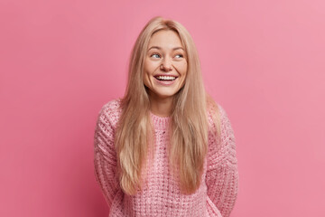 Wall Mural - Overjoyed young blonde woman smiles broadly has happy mood recalls funny moment happened with her dressed in knitted sweater isolated over pink background. People and sincere emotions concept
