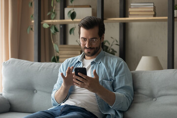 Wall Mural - young caucasian man in glasses sit rest on sofa at home look at cellphone screen texting or messagin