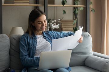 Happy millennial Caucasian female sit on couch study distant on laptop at home, read paperwork considering. Smiling young woman work with paper documents or correspondence work online on computer.